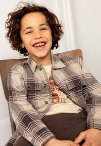 Geruite shirt in beige, bruin en blauw met drukknoopjes en twee borstzakken, gedragen over een lichtbeige t-shirt met opdruk en bruine broek.