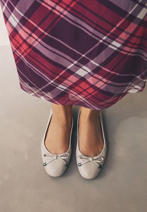 Jalad heledates beežides baleriinades ja lilla-punase ruudulise seelikuga, mis on pahkluude kohal nähtav neutraalsel põrandal.