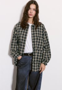 Chemise en flanelle à carreaux bleu marine et beige avec une coupe décontractée, dotée d'un bouton sur le devant, de manches longues et d'une poche poitrine. Portée par-dessus un t-shirt blanc.