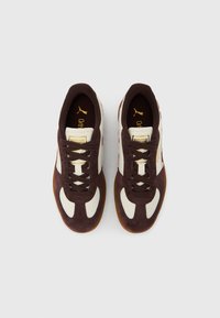 Barna és fehér Puma tornacipő pár, fűzőkkel és ortopéd talpbetétekkel, felülről nézve, egyszerű háttéren.