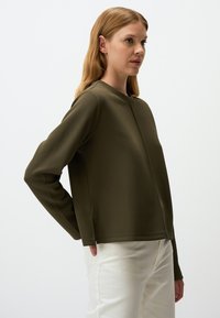 Felpa verde oliva a maniche lunghe con scollo rotondo, materiale testurizzato, spacchi laterali e vestibilità comoda; abbinata a pantaloni chiari.