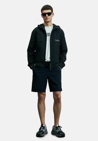 Veste noire à capuche avec fermeture éclair, associée à un t-shirt blanc et à un short noir. Portée avec des lunettes de soleil noires et des chaussures de sport.