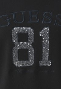 Svart tyg med ett mönster där "GUESS" är broderat ovanför siffran "81", prytt med silverpaljetter och en skrivstil nedanför.