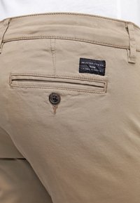 Béžové bavlněné kalhoty s zadní kapsou, zapínáním na knoflík a černou štítkem s nápisem "SELECTED HOMME" při švu v pase.