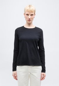 Άτομο με κοντά ξανθά μαλλιά που φοράει μαύρο μακρυμάνικο πουκάμισο και λευκό παντελόνι, στέκεται μπροστά σε απλό ανοιχτό φόντο.