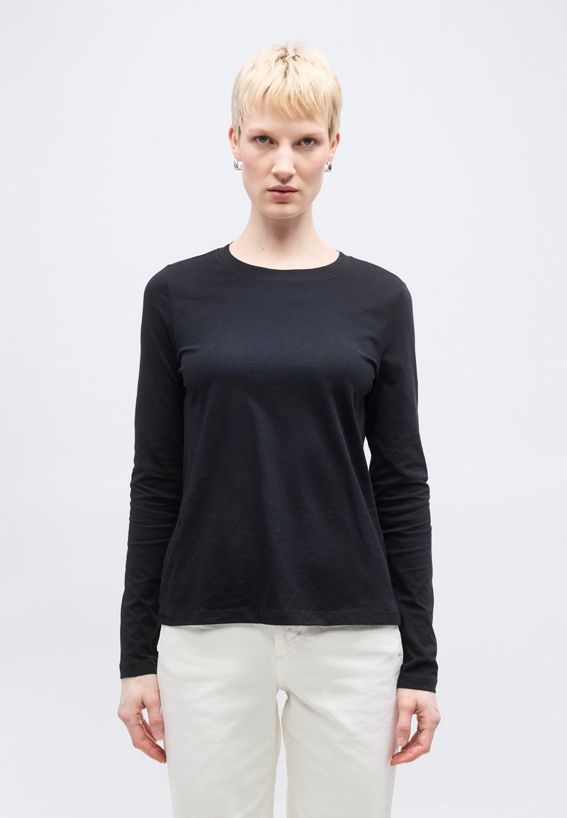 Άτομο με κοντά ξανθά μαλλιά που φοράει μαύρο μακρυμάνικο πουκάμισο και λευκό παντελόνι, στέκεται μπροστά σε απλό ανοιχτό φόντο.