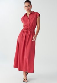 Rød maxikjole med korte ærmer, knaplukning foran, snore i taljen, glat tekstur og afslappet pasform. Bæres med sorte sandaler.