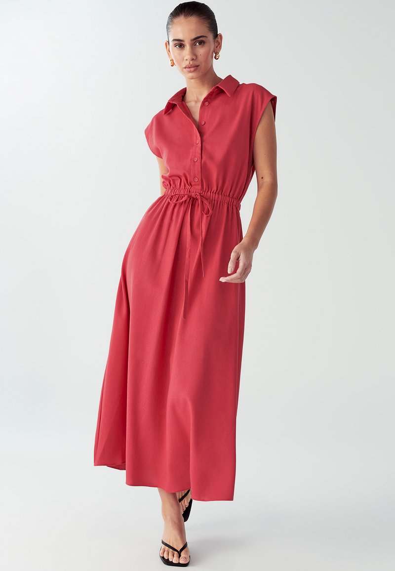 Rød maxikjole med korte ærmer, knaplukning foran, snore i taljen, glat tekstur og afslappet pasform. Bæres med sorte sandaler.