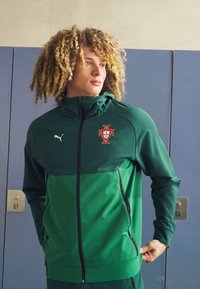 Zaļa zip-up jaka ar kontrastējošu augšējo paneli, ar Portugāles emblemu un Puma logo. Gluda tekstūra, sportiska piegriešana un praktiskas kabatas ar zipu.
