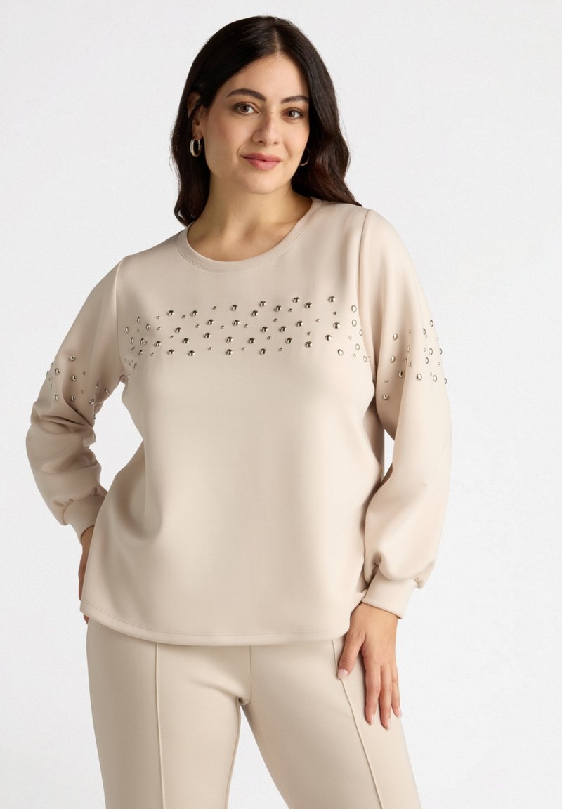 Beige long-sleeve top featuring embellished details on sleeves and chest, smooth texture, rounded neckline, and relaxed fit.