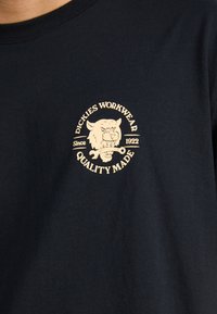Juodas medvilninis marškinėliai su apvaliu, kreminės spalvos logotipu, kuriame pavaizduotas liūto veidas, tekstas "DICKIES WORKWEAR" ir "QUALITY MADE".