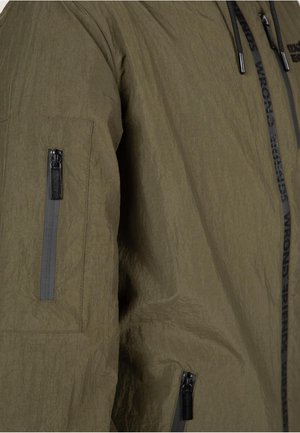 Olive green jacket with a textured finish, featuring a zippered pocket on the sleeve and a logoed strap along the zipper.