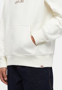 Witte sweatshirt met een buidelzak, ribgebreide manchetten en zoom. "Quality Made" geborduurd in bruin. Gecombineerd met een donkere spijkerbroek.