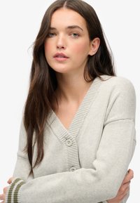 Jasnoszary kardigan z dekoltem w serek, prążkowanymi detalami i dwoma guzikami. Mankiety ozdobione zielono-białym paskiem. Miękka tekstura.