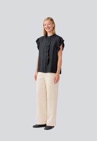 Black short-sleeve blouse with ruffle detailing and button closure, paired with textured cream wide-leg pants and black shoes.