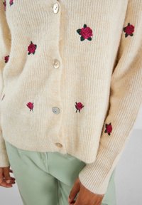 Beige kavaj i mjuk ribbad tyg, med små broderade rosor i röd och grön, prydd med pärlliknande knappar.