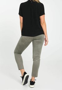 Femme se tenant sur une jambe pliée, portant un haut noir à manches courtes, un pantalon gris slim et des baskets noires à semelles blanches.