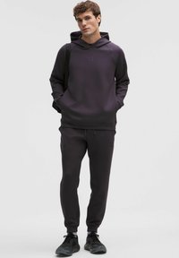 Mørkegrå hættetrøje med kængurulomme og matchende joggers. Materialet virker blødt. Har logo på brystet, og skoene er sorte med detaljer.