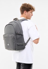 Un tânăr îmbrăcat cu un rucsac mare gri, purtând un tricou alb, într-un fundal alb simplu, așezat de profil.