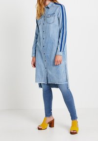 Denimmekko, jossa on napitus, pitkät hihat ja kaksi etutaskua; siinä on siniset raidalliset yksityiskohdat hihoissa ja repaleinen helma. Yhdistetty keltaisiin korkokenkiin.