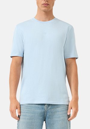 Mężczyzna ubrany w prostą jasnoniebieską koszulkę z krótkim rękawem i jasnoniebieskie dżinsy, stojący na jednolitym tle.