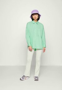 Jovem mulher vestindo uma camisa oversized verde menta, calças brancas caneladas, tênis cinzas e um chapéu tipo balde lavanda, contra um fundo liso.