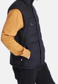 Czarna puchowa kamizelka z wysokim kołnierzem i zapięciem na zatrzaski, z bocznymi kieszeniami. Nosi się ją na musztardowej bluzie z ściągaczami przy mankietach.