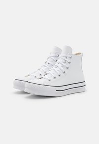 Zapatillas altas blancas de lona, con punta redonda, franja negra en la suela, ojales metálicos y cordones planos blancos.