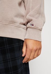 Sudadera beige con puños de canalé, que presenta una pequeña etiqueta de logo en rojo, blanco y azul. Lleva puesta sobre una falda de cuadros oscuros.