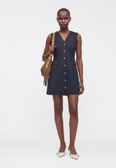 Femme portant une robe sans manches bleu marine à fines rayures avec des boutons, tenant un sac à bandoulière marron à franges, et chaussée de souliers blancs à bout pointu avec des nœuds.