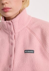 Close-up van een persoon die een roze Columbia fleece jack draagt met drukknoopjes en een hoge kraag.