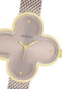 Montre à quartz en forme de trèfle à quatre feuilles, avec un cadran gris, des accents dorés et un bracelet en maille argentée. Les caractéristiques incluent des aiguilles des heures et des minutes simples.