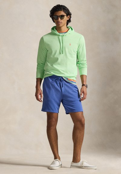 Homme portant un sweat à capuche vert, une chemise rayée, un short bleu, des baskets blanches, des lunettes de soleil et une montre-bracelet, debout devant un fond neutre.