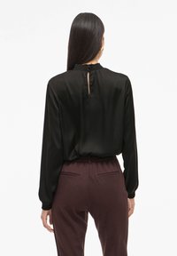 Schwarze Satin-Bluse mit langen Ärmeln, hohem Kragen, Raffungen und einer Schlitzöffnung am Rücken. Kombiniert mit bordeauxroten hochgeschnittenen Hosen.