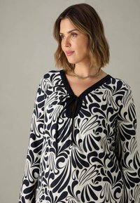 Svartvit mönstrad blus med V-ringning, knyt-detalj vid halsen och lång ärm. Tillverkad av ett slätt, lätt material.