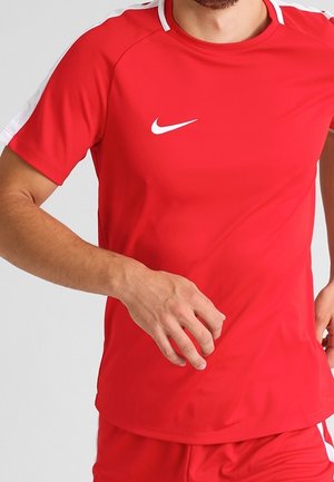 Mężczyzna w czerwonej sportowej koszulce i krótkich spodenkach Nike z białymi akcentami, w ruchu z prawą ręką w pobliżu talii.