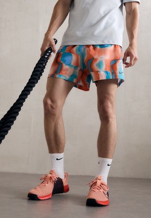 Mężczyzna w kolorowych sportowych szortach, białych skarpetkach Nike, koralowych butach sportowych Nike, trzymający czarną linę treningową na jednolitym tle.