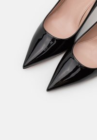 Escarpins noirs en cuir verni à bouts pointus avec doublure intérieure beige, posés sur un fond blanc.