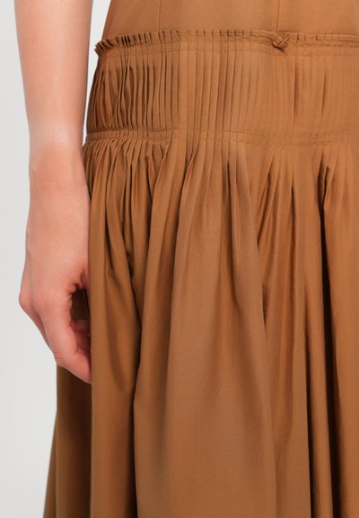 Braunes Stoffkleid mit geraffter Taille und plissiertem Rock. Der Stoff ist glatt und erzeugt eine fließende Textur. Nahaufnahme der Kleiderdetails und einer Hand.