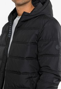 Czarna kurtka puchowa z kapturem, pikowanym wzorem, zapięciem na zamek i bocznymi kieszeniami. Materiał wydaje się gładki z delikatnym połyskiem.