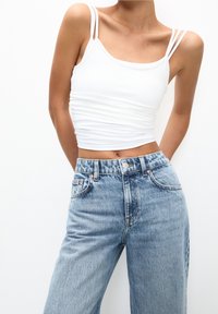 Weißes Crop-Top mit dünnen Trägern, kombiniert mit hoch geschnittenen hellblauen Jeans. Glatte Textur, lässige Passform und keine sichtbaren Muster oder Akzente.
