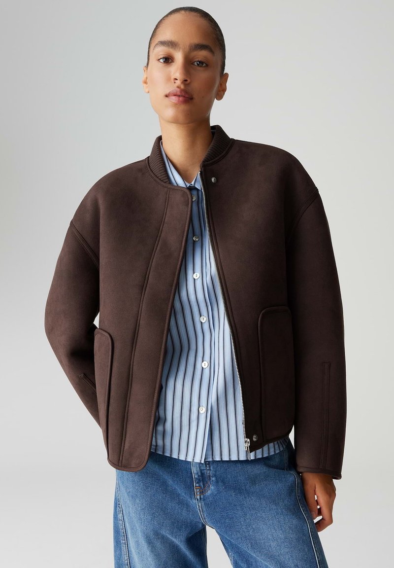 Giacca marrone oversize con texture liscia, colletto a costine e tasche laterali, indossata sopra una camicia a righe blu e bianche con bottoni.