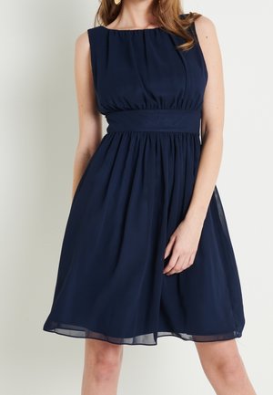 Frau trägt ärmelloses, marineblaues knieumspielendes Kleid mit geraffter Taille und fließendem Rock vor einfachem Hintergrund.