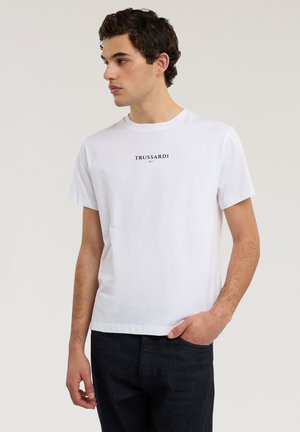 Młody mężczyzna w białej koszulce Trussardi z okrągłym dekoltem i ciemnych dżinsach, patrzący w lewo, z jedną ręką w kieszeni, na jednolitym tle.