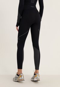 Mustat urheilulegginsit korkealla vyötäröllä, joissa on heijastavat harmaat yksityiskohdat ja verkkopaneelit alaosassa. Yhdistetty juoksukenkiin.