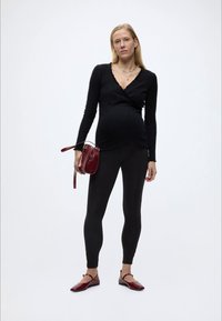 Gravid kvinne iført svart langermet topp og leggings, holder en burgunder håndveske, med matchende burgunder ballerinaer, stående mot hvit bakgrunn.