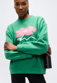 Γυναίκα με πράσινο φούτερ με σχέδιο ροζ λουλουδιού, σταυρωμένα χέρια, κρατώντας μαύρο μπουκάλι νερού, κοιτάζοντας μπροστά σε απλό φόντο.