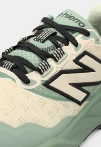 Light beige sneaker with mint green accents and a textured upper. Features black laces and a prominent black "N" logo on the side.