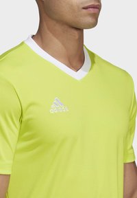 Lys gul idretts skjorte med hvit V-hals, med et brodert Adidas-logo på venstre bryst. Glatt, lett materiale.