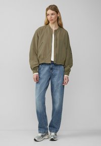 Olijfgroene bomberjack met geribbelde manchetten en tailleband, gecombineerd met lichtblauwe rechte spijkerbroek en zilveren sportschoenen.